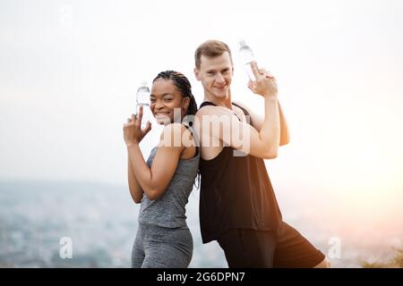 Porträt einer sportlichen afrikanischen Frau und eines kaukasischen Mannes, der mit einer Flasche Wasser in den Händen wie eine Waffe zurücksteht und vor der Kamera lächelt. Junges Paar, das aktiv an der frischen Luft seine Freizeit verbringt. Stockfoto