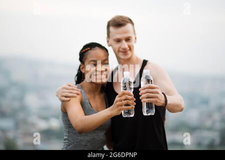 Positives multirassisches Paar in aktiver Kleidung, das nach dem Training im Freien mit einer Flasche Wasser toaste. Konzept von Menschen, Sport und Erholung. Stockfoto