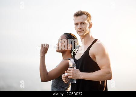 Schöner kaukasischer Mann und schöne afrikanische Frau halten eine Flasche Wasser, während sie im Freien stehen. Sportliches junges Paar im aktiven Ohr, das nach dem Training den Wasserhaushalt erneuert. Stockfoto