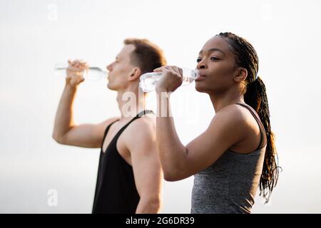Seitenansicht einer afroamerikanischen Frau und eines kaukasischen Mannes, der sich nach einem aktiven Training im Freien mit kaltem Wasser erfrischt. Konzept der Menschen, gesunde Lebensweise und Erholung. Stockfoto