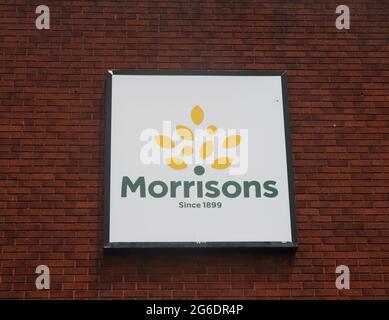 Frontaler Nahaufnahme des quadratischen Morrisons-Logos mit gelben Spritzern, Markenname und Since 1899-Text auf einem dunkelroten Ziegelwandhintergrund. Stockfoto