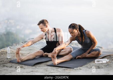 afroamerikanische Frau und kaukasischer Mann strecken sich die Beine auf einer Yogamatte auf hohem Felsen. Junges Paar in aktivem Ohr, das gemeinsam an der frischen Luft trainiert. Stockfoto