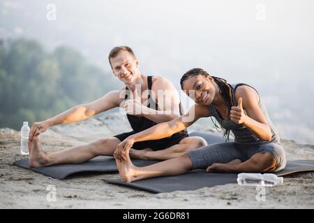 afroamerikanische Frau und kaukasischer Mann strecken sich die Beine auf einer Yogamatte auf hohem Felsen. Junges Paar in aktivem Ohr, das gemeinsam an der frischen Luft trainiert. Stockfoto