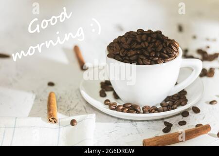 Weiße Tasse mit Kaffeebohnen und dem Text Guten Morgen. Guten Morgen geschrieben. Kaffeebohnen in einer Kaffeetasse und Zimtstangen. Hintergrund mit Text Stockfoto