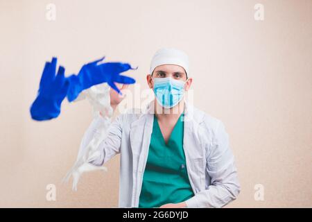 Junge Arzt Porträt in weißen Laborkittel schützende Atemmaske wirft weiße und blaue Handschuhe auf gelben Hintergrund isoliert. Stockfoto