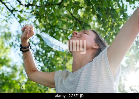 Glückliche Frau wird am Sommertag eine medizinische Maske aus ihrem Gesicht entfernen. Konzept der Bekämpfung des Virus, das Ende der Pandemie. Stockfoto