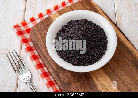 Rohe, trockene schwarze Reiskörner in einer weißen Keramikschale auf einem braunem Holzschneidebrett über einem Holztisch. Unverarbeiteter schwarzer Bio-Reis für die Ernährung Stockfoto