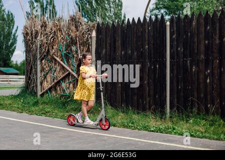 Ein Spaziergang auf einem Roller eines Mädchens in gelbem Kleid an einem Holz- oder Schilfzaun an einem Sommertag Stockfoto