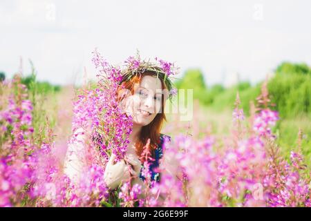 Porträt einer jungen, schönen rothaarigen Frau mit einem Kranz auf ihrem Kopf. Lächelnde Dame steht auf einem Feld mit wilden rosa Blumen. Stockfoto
