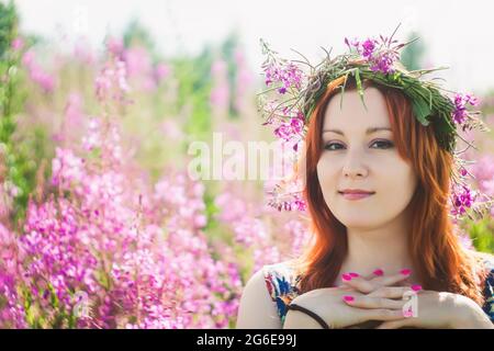 Die junge, wunderschöne rothaarige Frau mit einem Kranz rosa Wildblumen auf ihrem Kopf blickt auf die Kamera und lächelt. Sommerlicher Style Stockfoto