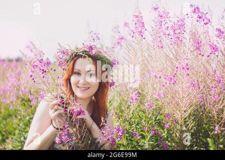 Porträt einer jungen, schönen rothaarigen Frau mit einem Kranz auf ihrem Kopf. Lächelnde Dame steht auf einem Feld mit wilden rosa Blumen. Stockfoto