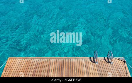 Yacht Heck Deck, Teakholz und Metall Leitern, türkisblaues Meer Wasser Hintergrund. Segelboot liegt am offenen Meer. Schwimmen in der Ägäis, Greec Stockfoto