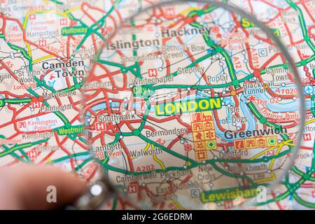 Nahaufnahme einer Seite in einem gedruckten Straßenkarte-Atlas mit einer Männerhand, die eine Lupe mit einer Vergrößerung von London in England hält Stockfoto