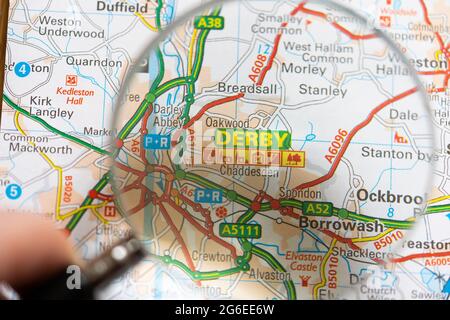 Nahaufnahme einer Seite in einem gedruckten Straßenkarte-Atlas mit einer Männerhand, die eine Lupe hält, die eine Vergrößerung der Stadt Derby, England, zeigt Stockfoto