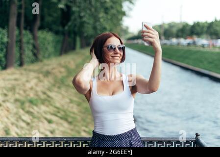 Das rothaarige Mädchen lacht in einer Sonnenbrille und macht im Sommer im Park ein Selfie auf einem Smartphone. Stockfoto