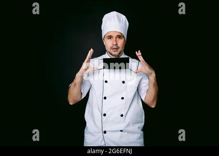 Der Koch trägt eine Kochjacke und einen Hut, setzt eine schwarze Maske auf schwarzem Hintergrund auf Stockfoto