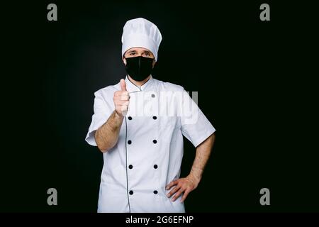 Chefkoch trägt Kochjacke und Hut, mit schwarzer Maske, Daumen heben, auf schwarzem Hintergrund Stockfoto