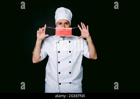 Der Koch trägt eine Kochjacke und einen Hut, setzt eine rote Maske auf schwarzen Hintergrund auf Stockfoto