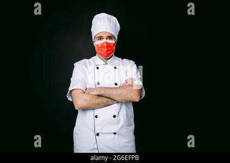 Koch trägt Kochjacke und Hut, mit roter Maske, gekreuzte Arme, auf schwarzem Hintergrund Stockfoto