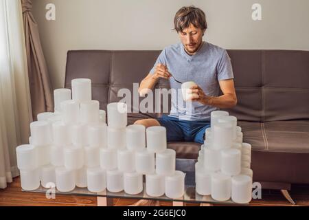 Der Mann isst Joghurt und ist von vielen Joghurtgläsern umgeben, die er in einem Jahr verwendet. Eine riesige Menge von Plastikbehältern für Lebensmittel. Übermäßiger Verbrauch von Stockfoto