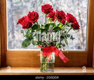 Hübsche rote Rosen in einer Glasvase mit einer roten Schleife vor einem Fenster mit Blick auf den Schnee draußen. Stockfoto