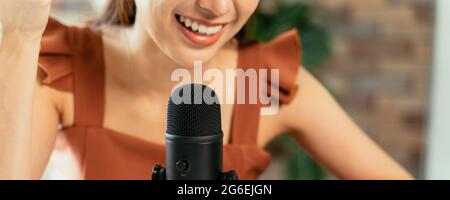 Nahaufnahme einer jungen, schönen Frau, die lächelnd auf dem Schreibtisch sitzt und dabei einen Podcast mit Laptop und Mikrofon aufzeichnet, während sie ein Interview führt Stockfoto