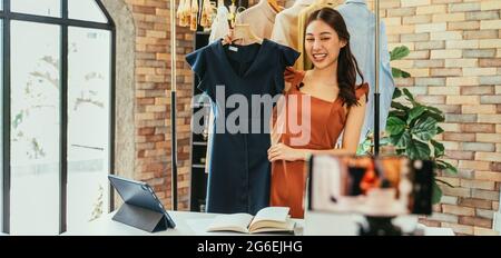 Junge und schöne asiatische Bloggerin zeigt Kleidung vor der Smartphone-Kamera, während sie Vlog-Video und Live-Streaming in ihrem Shop aufnimmt Stockfoto