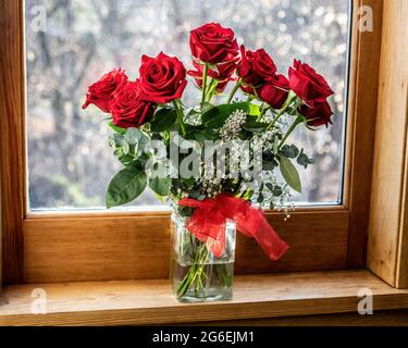 Hübsche rote Rosen in einer Glasvase mit einer roten Schleife vor einem Fenster mit Blick auf den Schnee draußen. Stockfoto