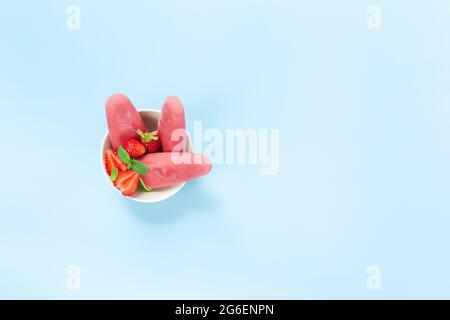 Draufsicht auf Schüssel mit fruchtigem, rosafarbenem, gefrorenem Sorbet-Eis auf Plastikstock mit Erdbeere, Minze auf blauem Hintergrund. Gefrorenes gemischtes Obst mit Joghurt i Stockfoto