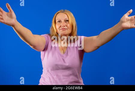 Mittelblonde Frau, die über blauem, isoliertem Hintergrund steht und die Kamera lächelt, während sie die offenen Arme umarmt. Fröhlicher Ausdruck umarmt happine Stockfoto