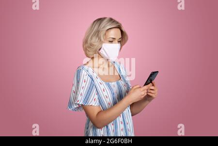 Die kaukasische Frau plaudert auf dem Handy, während sie eine medizinische Maske an einer roten Studiowand trägt Stockfoto