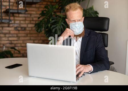 Nachdenklicher rothaariger Geschäftsmann in schickem Freizeitanzug, der eine schützende Gesichtsmaske trägt, mit einem Laptop, der im modernen Büro sitzt, schaut weg und sorgt sich um die Finanzkrise aufgrund einer Pandemie Stockfoto