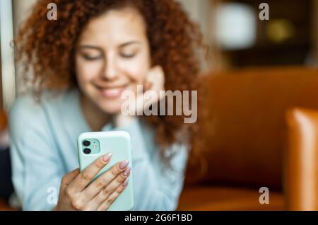Selektiver Fokus auf das trendige Smartphone in der Hand einer attraktiven rothaarigen lockigen Frau, einer Frau, die zu Hause telefonieren, eine Nachricht schreiben, online chatten Stockfoto
