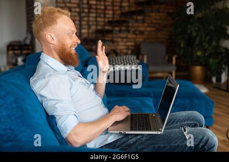 Lächelnder rothaariger Mann blickt auf die Kamera und winkt mit der Hand, Video-Chat mit bärtigen Hipster-Typen in elegantem Casual-Shirt. Videoanruf, virtuelles Meeting Stockfoto