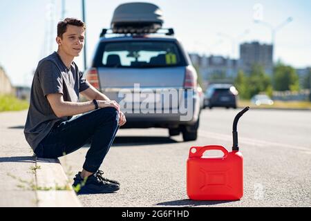 Ein roter Kanister auf dem Asphalt in der Nähe des Autos. Das Auto hatte kein Benzin mehr und war zum Stillstand gekommen. Ein junger Mann, der auf Hilfe von anderen Fahrern hofft Stockfoto