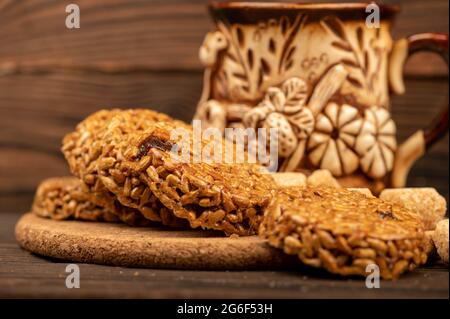 Hausgemachte Kekse mit Sonnenblumenkernen und Rosinen, Stücke von braunem Rohrzucker und ein Tonbecher Tee aus Steingut auf einem Holztisch. Nahaufnahme des selektiven Fokus Stockfoto