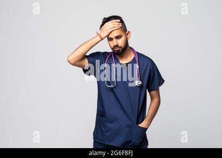 Porträt eines erschöpften indischen jungen Arztes mit Stethoskop in Uniform, der unter starken Kopfschmerzen leidet und isoliert auf die Kamera schaut Stockfoto
