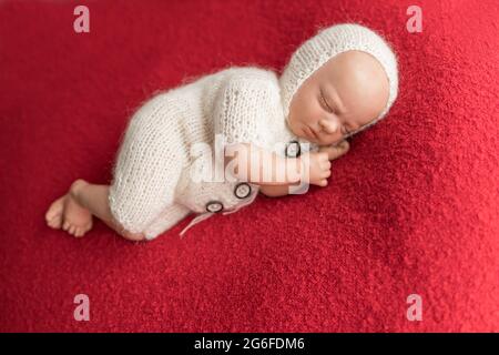 Die neugeborene Puppe trägt einen weißen Strickjumpsuit und eine Strickmütze, die auf ihrer Seite auf einer roten Decke liegt. Foto für Babys erste weihnachten, kein Modell r Stockfoto