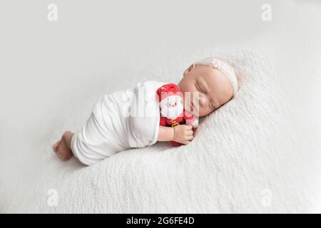 Die neugeborene Puppe, in einem weißen Wrap gewickelt, trägt ein weißes Perlkopfband und kuschelt einen weihnachtsmann, der auf ihrer Seite auf einer weißen Decke liegt. Stockfoto