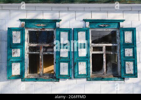 Zwei blaue Vintage-Fenster mit Holzrahmen, Fensterläden und abblätternder Farbe mit Kratzern an der weißen Ziegelwand des alten Dorfhauses Stockfoto