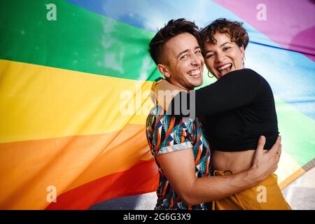 Lächelndes, queeres Paar, das gegen eine Regenbogenflagge steht. Junge LGBTQ-Paare umarmen sich gegenseitig, während sie zusammen stehen. Zwei nicht konforme Liebhaber Stockfoto