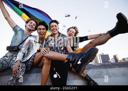 Freunde feiern schwulen Stolz, während sie zusammen sitzen. Vier Mitglieder der LGBTQ+ Community lächeln fröhlich und heben die Pride-Flagge. Gruppe von q Stockfoto