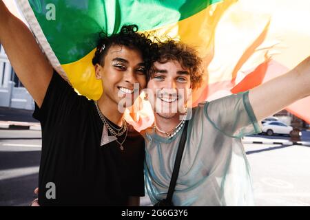 Zwei junge, schwule Männer feiern den Stolz der Homosexuellen. Zwei junge schwule Männer lächeln fröhlich, während sie bei einer Parade zum Stolz der Homosexuellen eine Regenbogenfahne heben. Zwei Mitglieder des Stockfoto