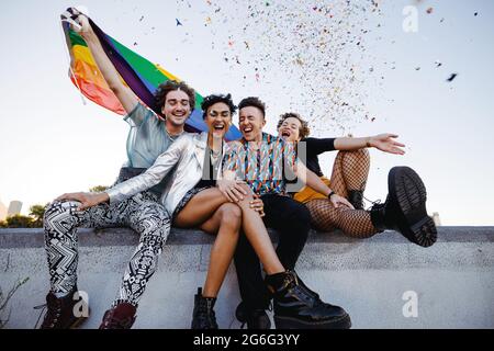 Fröhliche, queere Menschen feiern den Stolz der Homosexuellen. Gruppe von vier Freunden, die die Regenbogenflagge halten, während sie gemeinsam im Freien sitzen. Fröhliche Mitglieder von Th Stockfoto