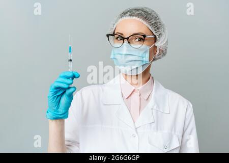 Ärztin oder Wissenschaftlerin in Weiß medizinische Kleid, blau Handschuhe, grüne Kappe und Maske hält eine Spritze in der Hand auf weißem Hintergrund. Sie ist bereit zu geben Stockfoto