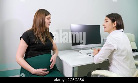 Aufmerksamer Brünette Arzt in weißem Gewand konsultiert junge langhaarige Schwangere, die mit einem modernen Computer am Tisch in einem geräumigen Klinikbüro sitzt Stockfoto