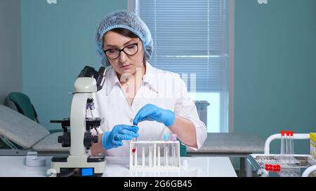 Frau Laborassistentin mit Brille und Kappe macht Tests unter Röhrchen mit verschiedenen Flüssigkeiten auf Tisch mit modernem Mikroskop in der Klinik Stockfoto