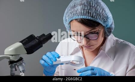 Ein konzentrierter Wissenschaftler mit Brille betrachtet den Blutstropfen auf dem Objektträger durch Lupenglas in der Nähe des Mikroskops, der in der Nahaufnahme des Krankenhauslabors arbeitet Stockfoto