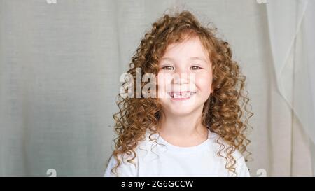 Lockiges rotbraunes Mädchen im Vorschulalter in weißem T-Shirt sieht gerade aus und lächelt mit verlorenen Zähnen, die für die Kamera auf grauem Hintergrund in Nahaufnahme posieren Stockfoto