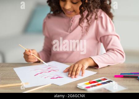 Inspiriertes afroamerikanisches Teenager-Mädchen mit Pinsel- und Palettenbemalung, das ihr neues Hobby genießt und zu Hause am Schreibtisch sitzt Stockfoto